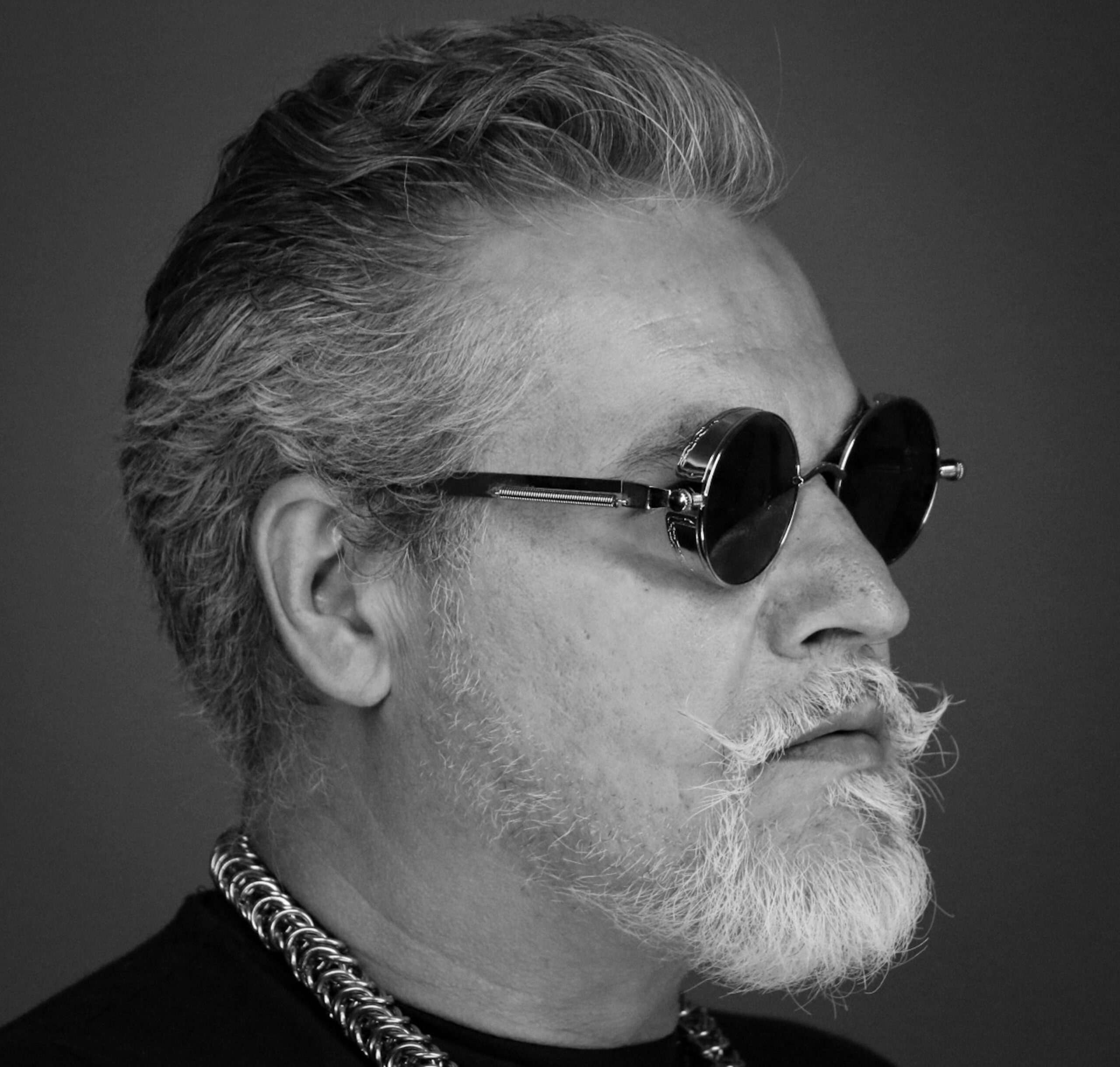 Stylish black and white headshot of an older man wearing sunglasses and a chain necklace.